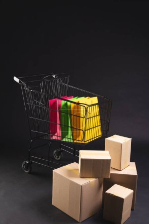 Photo for Vertical image of shopping trolley with bags, boxes and copy space over black background. Cyber monday, black friday, online shopping, shipping and global connections concept. - Royalty Free Image