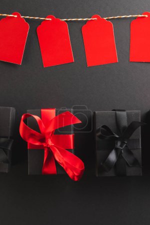 Photo for Vertical image of red gift tags on pegs and black gift boxes with copy space over black background. Cyber monday, black friday, online shopping, shipping and global connections concept. - Royalty Free Image