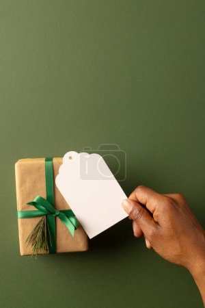 Photo for Vertical image of african american woman's hand holidng tag with copy space and christmas present. Presents, christmas, tradition and celebration concept. - Royalty Free Image