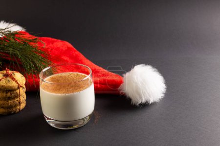 Téléchargez les photos : Verre de lait, biscuits de Noël et chapeau de Père Noël avec espace de copie sur fond noir. Bonbons, Noël, concept de tradition et de célébration. - en image libre de droit