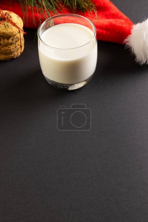 Photo for Vertical image of glass of milk, christmas cookies and santa hat with copy space on black background. Sweets, christmas, tradition and celebration concept. - Royalty Free Image