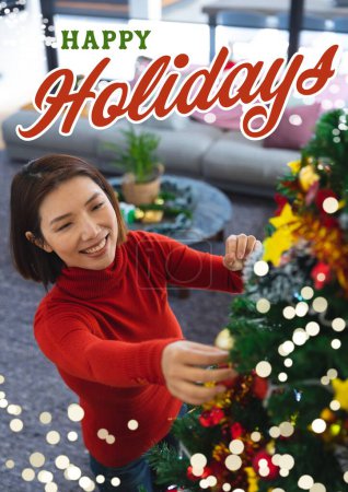Téléchargez les photos : Joyeux texte de vacances en vert et rouge sur une femme asiatique souriante décorant l'arbre de Noël. Noël, carte de voeux saisonniers, tradition et célébration image générée numériquement. - en image libre de droit