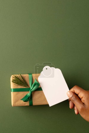 Photo for Vertical image of african american woman's hand holidng tag with copy space and christmas present. Presents, christmas, tradition and celebration concept. - Royalty Free Image
