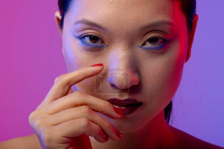 Foto de Retrato de mujer asiática con delineador de ojos azul y esmalte de uñas rojo sobre fondo púrpura. Cosméticos, maquillaje, moda femenina y belleza, inalterados. - Imagen libre de derechos