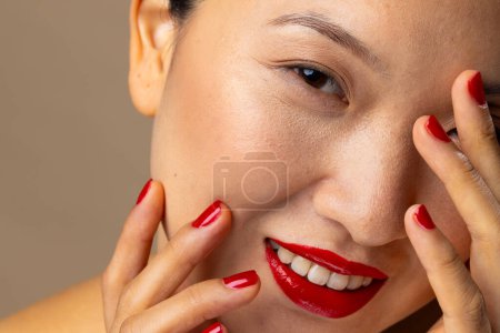 Foto de Retrato de mujer asiática con lápiz labial rojo y esmalte de uñas sobre fondo beige. Cosméticos, maquillaje, moda femenina y belleza, inalterados. - Imagen libre de derechos
