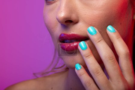 Foto de Mujer caucásica con lápiz labial rosa y esmalte de uñas azul sobre fondo púrpura. Cosméticos, maquillaje, moda femenina y belleza, inalterados. - Imagen libre de derechos