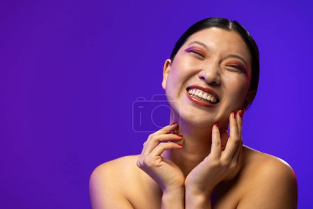Foto de Mujer asiática con sombra de ojos púrpura y esmalte de uñas rojo sobre fondo azul, espacio para copiar. Cosméticos, maquillaje, moda femenina y belleza, inalterados. - Imagen libre de derechos