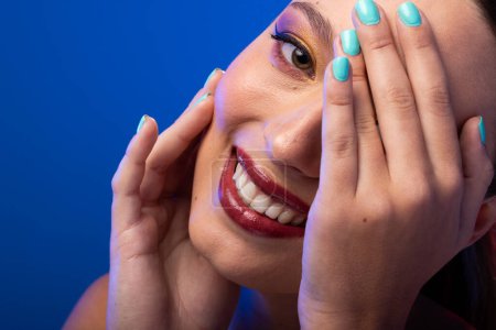 Téléchargez les photos : Portrait de femme caucasienne portant ombre à paupières dorée et vernis à ongles bleu sur fond bleu. Cosmétiques, maquillage, mode féminine et beauté, inchangés. - en image libre de droit