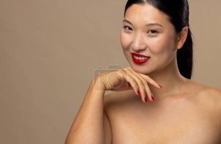 Foto de Retrato de mujer asiática con lápiz labial rojo y esmalte de uñas sobre fondo beige, espacio para copiar. Cosméticos, maquillaje, moda femenina y belleza, inalterados. - Imagen libre de derechos