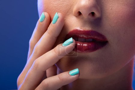 Foto de Mujer caucásica con lápiz labial rojo y esmalte de uñas azul sobre fondo azul. Cosméticos, maquillaje, moda femenina y belleza, inalterados. - Imagen libre de derechos