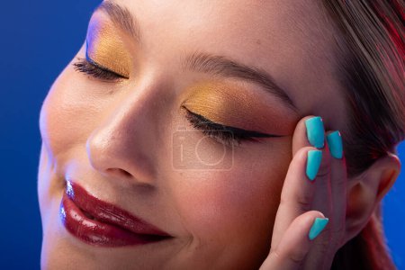 Foto de Mujer caucásica con cabello castaño con sombra de ojos dorada y esmalte de uñas azul sobre fondo azul. Cosméticos, maquillaje, moda femenina y belleza, inalterados. - Imagen libre de derechos