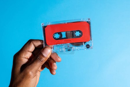 Foto de Primer plano de la mano del hombre afroamericano sosteniendo cinta de casete roja sobre fondo azul. Música, sonido, escucha, entretenimiento y concepto de nostalgia. - Imagen libre de derechos
