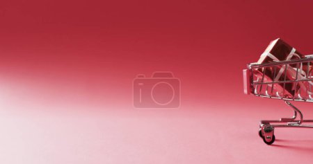 Photo for A miniature shopping cart holds a gift on a red background, with copy space. Symbolizes the convenience of shopping for presents and the spirit of giving. - Royalty Free Image
