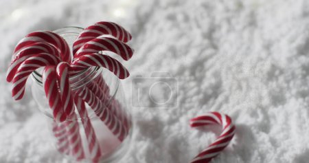 Foto de Los bastones de caramelo se derraman de un frasco sobre una superficie nevada. Evocando alegría navideña, las rayas rojas y blancas simbolizan dulzura festiva. - Imagen libre de derechos