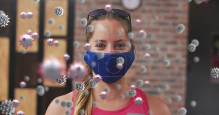 Foto de Múltiples células covid-19 flotando contra el retrato de una mujer caucásica en forma usando una máscara en el gimnasio. deportes, fitness y covid-19 concepto pandémico - Imagen libre de derechos