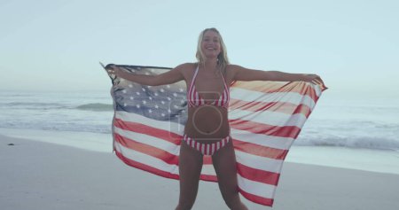 Téléchargez les photos : La jeune femme caucasienne se tient sur la plage tenant un drapeau américain. Elle porte un maillot patriotique symbolisant la fierté nationale au bord de l'océan.. - en image libre de droit