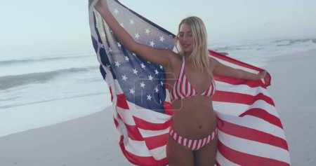 Téléchargez les photos : Happy 4th of july text against caucasian woman wrapping american around her on the beach. american independence patriotic holiday concept - en image libre de droit