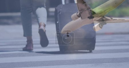 Photo for Airplane flying against low section of woman with trolley bag crossing the street. travel and vacation concept - Royalty Free Image