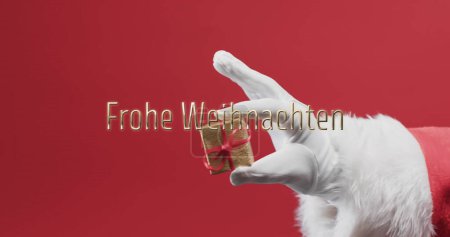 Foto de Frohe weihnachten texto sobre la mano de padre navidad sosteniendo pequeño regalo sobre fondo rojo. Navidad, alemán, tradición, saludos y celebración imagen generada digitalmente. - Imagen libre de derechos