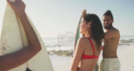 Photo for Young Caucasian woman and young Biracial man at the beach. They're enjoying a sunny day with a surfboard, ready for some ocean waves. - Royalty Free Image