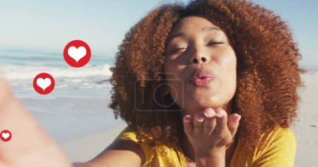 Foto de Imagen de corazón rojo amor iconos digitales sobre sonriente mujer soplando besos en la playa. interfaz digital, redes sociales y concepto de red global imagen generada digitalmente. - Imagen libre de derechos