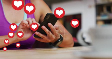 Téléchargez les photos : Plusieurs icônes cardiaques flottant contre la section médiane de la femme caucasienne à l'aide d'un smartphone. concept de technologie de réseaux sociaux - en image libre de droit