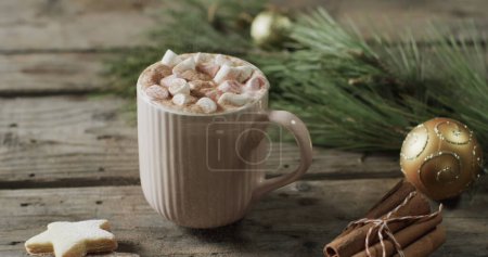 Photo for A mug of hot chocolate topped with marshmallows sits on a wooden table. Festive decorations suggest a cozy holiday atmosphere at home. - Royalty Free Image