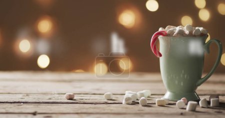Téléchargez les photos : Une tasse confortable de chocolat chaud ornée de guimauves sur une table en bois. Les lumières festives créent une atmosphère chaleureuse et accueillante, parfaite pour les vacances de détente. - en image libre de droit