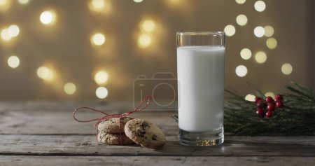 Foto de Un vaso de leche y galletas sobre una mesa de madera, con espacio para copiar. Luces festivas crean un ambiente cálido de vacaciones en el fondo. - Imagen libre de derechos