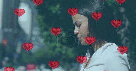 Foto de Mujer birracial rodeada de globos cardíacos, con espacio para copias. Ella está al aire libre, reflexionando sobre un momento romántico o celebración. - Imagen libre de derechos