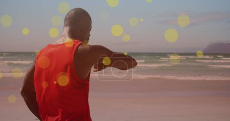 Foto de Manchas amarillas flotando contra el hombre afroamericano en forma usando smartwatch en la playa. concepto de deportes, fitness y tecnología - Imagen libre de derechos