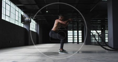 Téléchargez les photos : Jeune homme biracial effectue squats dans une salle de gym. Sa routine d'entraînement met en valeur la force et l'endurance dans un espace de fitness de style industriel. - en image libre de droit