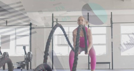 Foto de Imagen de los datos de la interfaz sobre el entrenamiento cruzado de mujeres caucásicas con cuerdas de batalla en el gimnasio. Fitness, ejercicio, fuerza, datos, interfaz digital y tecnología de imagen generada digitalmente. - Imagen libre de derechos