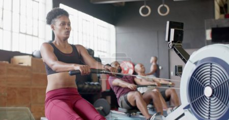 Foto de Joven mujer afroamericana hace ejercicios en un remo en el gimnasio. Los hombres biraciales participan en sus rutinas de entrenamiento en el fondo, manteniendo la aptitud. - Imagen libre de derechos