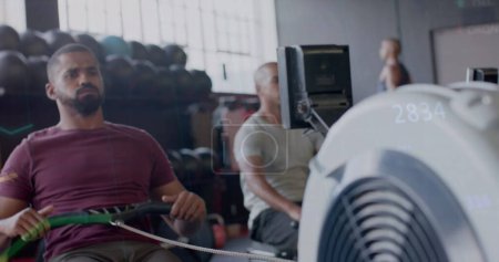 Foto de Imagen del procesamiento de datos en gráfico sobre diversos hombres entrenando en máquinas de remo en el gimnasio. Fitness, ejercicio, fuerza, datos, interfaz digital y tecnología de imagen generada digitalmente. - Imagen libre de derechos