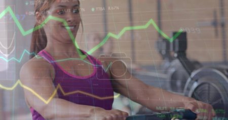 Foto de Imagen del procesamiento de datos en gráfico sobre el entrenamiento de la mujer birracial feliz en la máquina de remo en el gimnasio. Fitness, ejercicio, fuerza, datos, interfaz digital y tecnología de imagen generada digitalmente. - Imagen libre de derechos