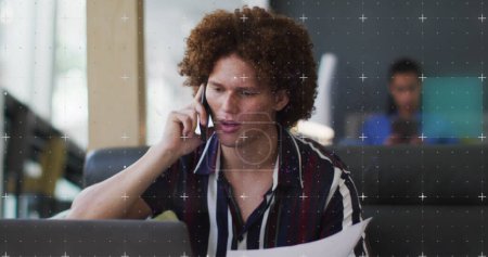 Un joven birracial habla por teléfono en una oficina. Se centra en una conversación mientras sostiene documentos, lo que indica un entorno empresarial.