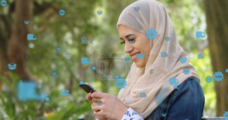 Foto de Red de iconos digitales contra la mujer en hijab sonriendo mientras usa el teléfono inteligente en el parque. concepto global de redes y tecnología - Imagen libre de derechos