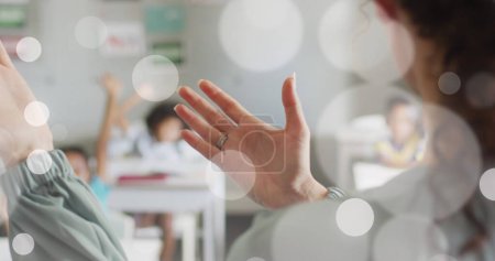 Image of white bokeh spots over hands of diverse female teacher and pupils in class. Communication, teaching, school, education, childhood and learning, digitally generated image.