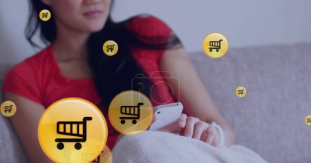 Téléchargez les photos : Chariots d'achat icônes flottant contre la femme asiatique en utilisant un smartphone sur le canapé à la maison. achats en ligne et concept de technologie - en image libre de droit
