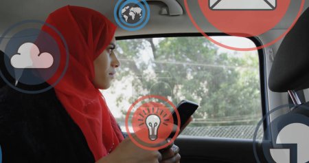 Foto de Múltiples iconos digitales flotando contra la mujer en hijab utilizando el teléfono inteligente en el coche. concepto global de redes y tecnología - Imagen libre de derechos