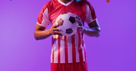 Foto de Hombre afroamericano sostiene una pelota de fútbol, con espacio para copiar. Atleta en un jersey rojo y blanco se levanta sobre un fondo púrpura. - Imagen libre de derechos