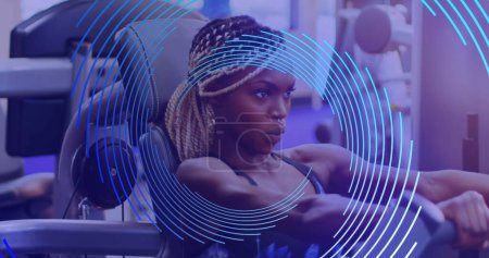 Téléchargez les photos : Image d'une spirale de ligne bleue tournant sur une femme afro-américaine faisant de l'exercice au gymnase. concept sport et compétition, image générée numériquement. - en image libre de droit