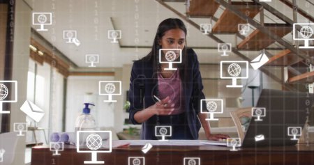 Image of icons and data processing over biracial businesswoman working in office. Global digital interface, cloud computing and data processing concept digitally generated image.