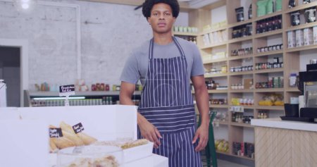 Téléchargez les photos : Le jeune Afro-Américain se tient en confiance dans une boulangerie, avec un espace de copie. Son tablier suggère qu'il est boulanger ou employé de magasin, entouré de pâtisseries fraîches. - en image libre de droit