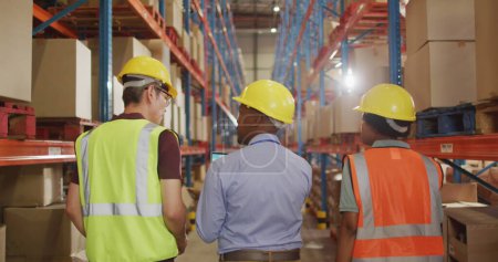 Equipe diversifiée de travailleurs d'entrepôt en discussion, avec espace de copie. Les casques et gilets de sécurité indiquent que l'accent est mis sur la sécurité au travail dans un cadre industriel.