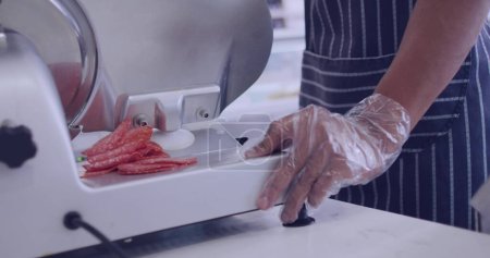 Person schneidet Salami auf einem Fleischschneider in einem Feinkostladen. Der Fokus auf die Zubereitung von Lebensmitteln legt eine professionelle Küche oder ein kulinarisches Umfeld nahe.