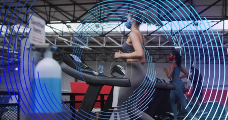 Téléchargez les photos : Image d'une spirale bleue tournant sur des femmes portant des masques sur des machines en marche. sport, fitness et technologie pendant une pandémie covid, image générée numériquement. - en image libre de droit