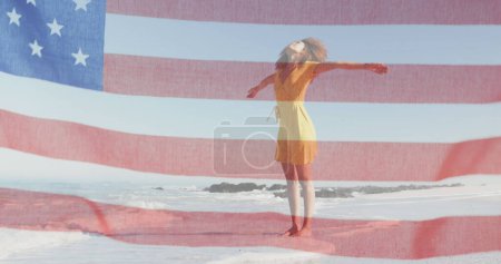 Foto de Bandera americana ondeando contra mujer afroamericana de pie con los brazos abiertos en la playa. - Imagen libre de derechos