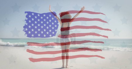 Photo for American flag waving over african american woman standing with arms wide open at the beach - Royalty Free Image